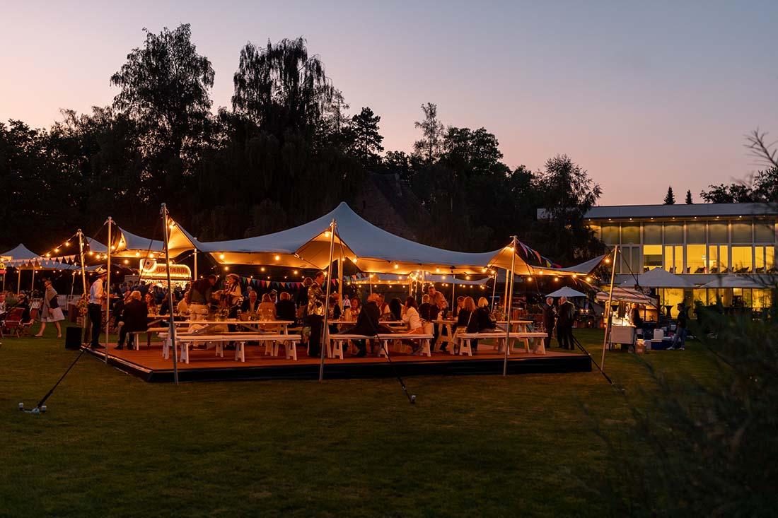 Ein grosses Eventzelt, das mit Gästen gefüllt ist, die unter dem sanften Glanz von Lichterketten speisen. Der Hintergrund zeigt den modernen GDI-Bau und einen dämmernden Himmel.